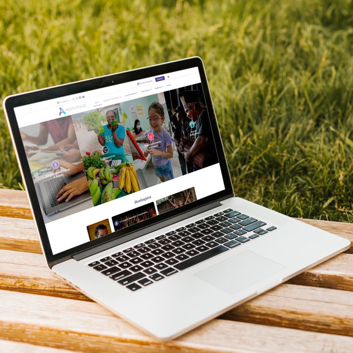 Laptop exibindo a página inicial do site do Instituto Alok, posicionado em uma mesa de madeira ao ar livre com grama verde ao fundo.